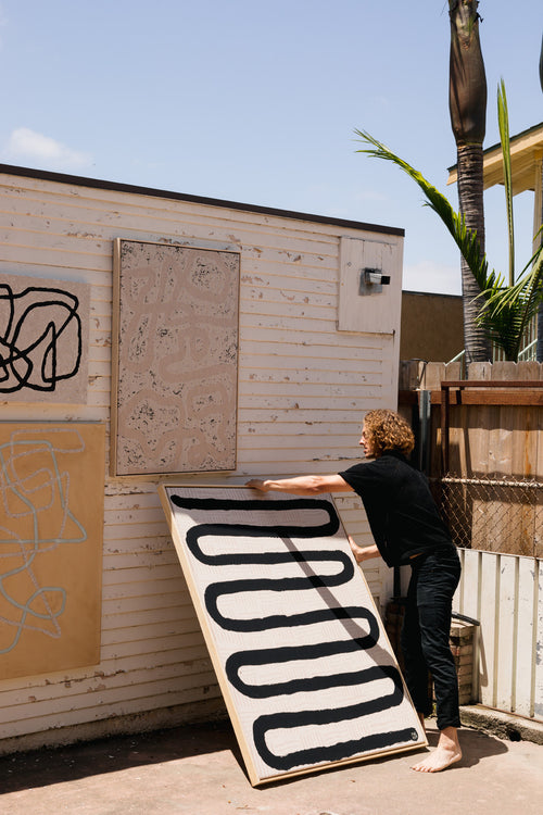 person moving large rectangular abstact art painting off of distressed wall, with other abstact art pieces, palm trees in background