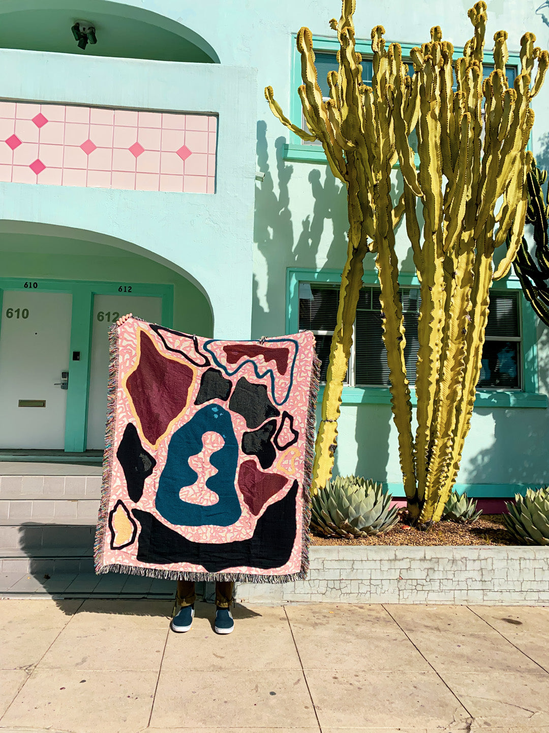 outdoor lifestyle shot of abstract art woven throw blanket being held up like a tapestry in front of bright green cactus, and lime green building with pink accents