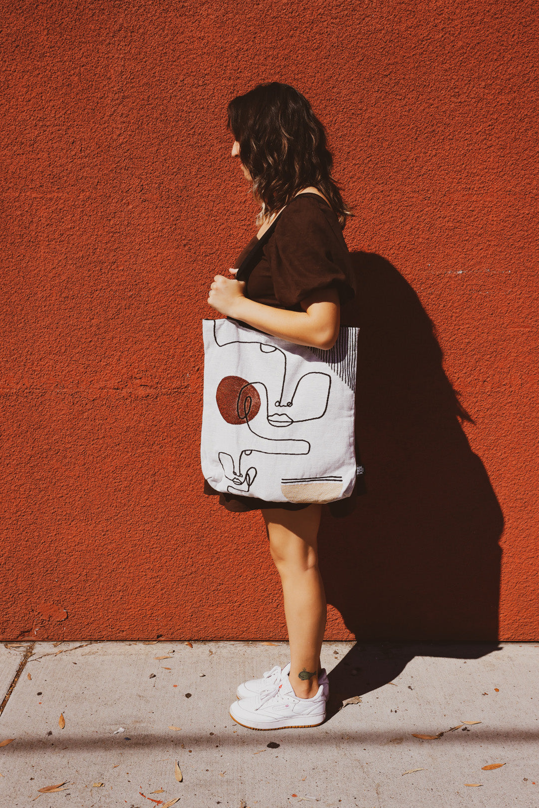 girl standing profile drenched in sunlight holding abstract art tote bag, with minimalist face design, bright bold burnt red in background